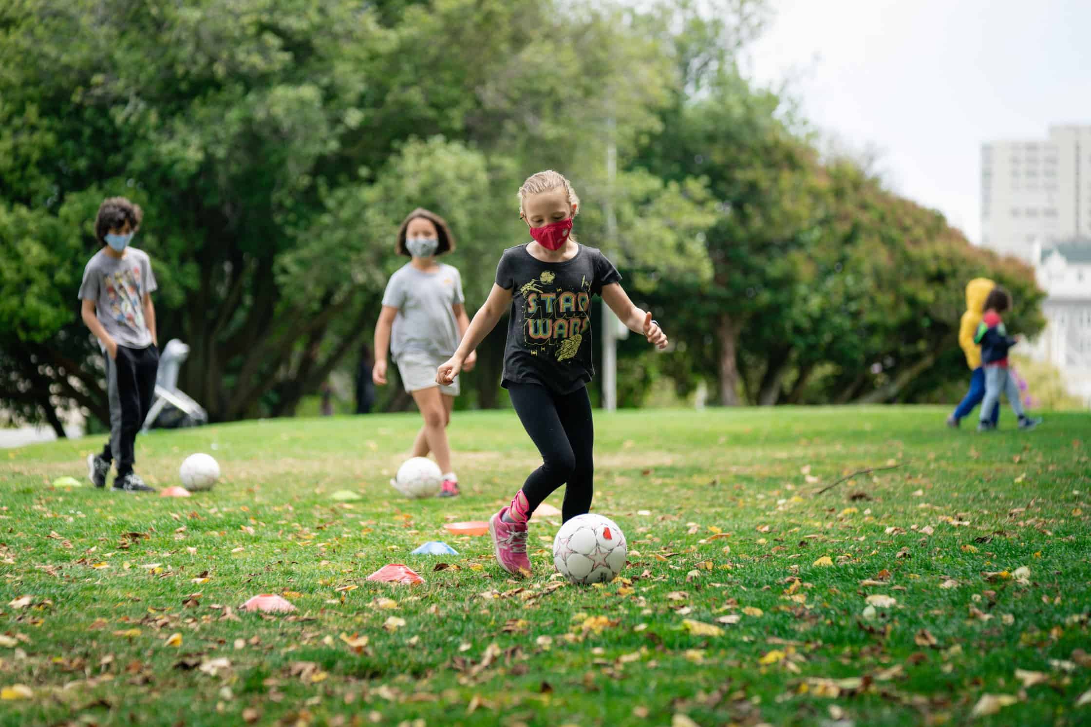 Elementary students play soccer in a field while wearing masks.